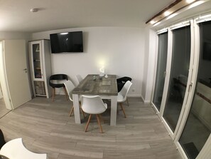 Ein Essbereich mit Holztisch und weißen Stühlen, einem an der Wand befestigten Flachbildfernseher und einer Glasschiebetür zu einem Außenbereich.