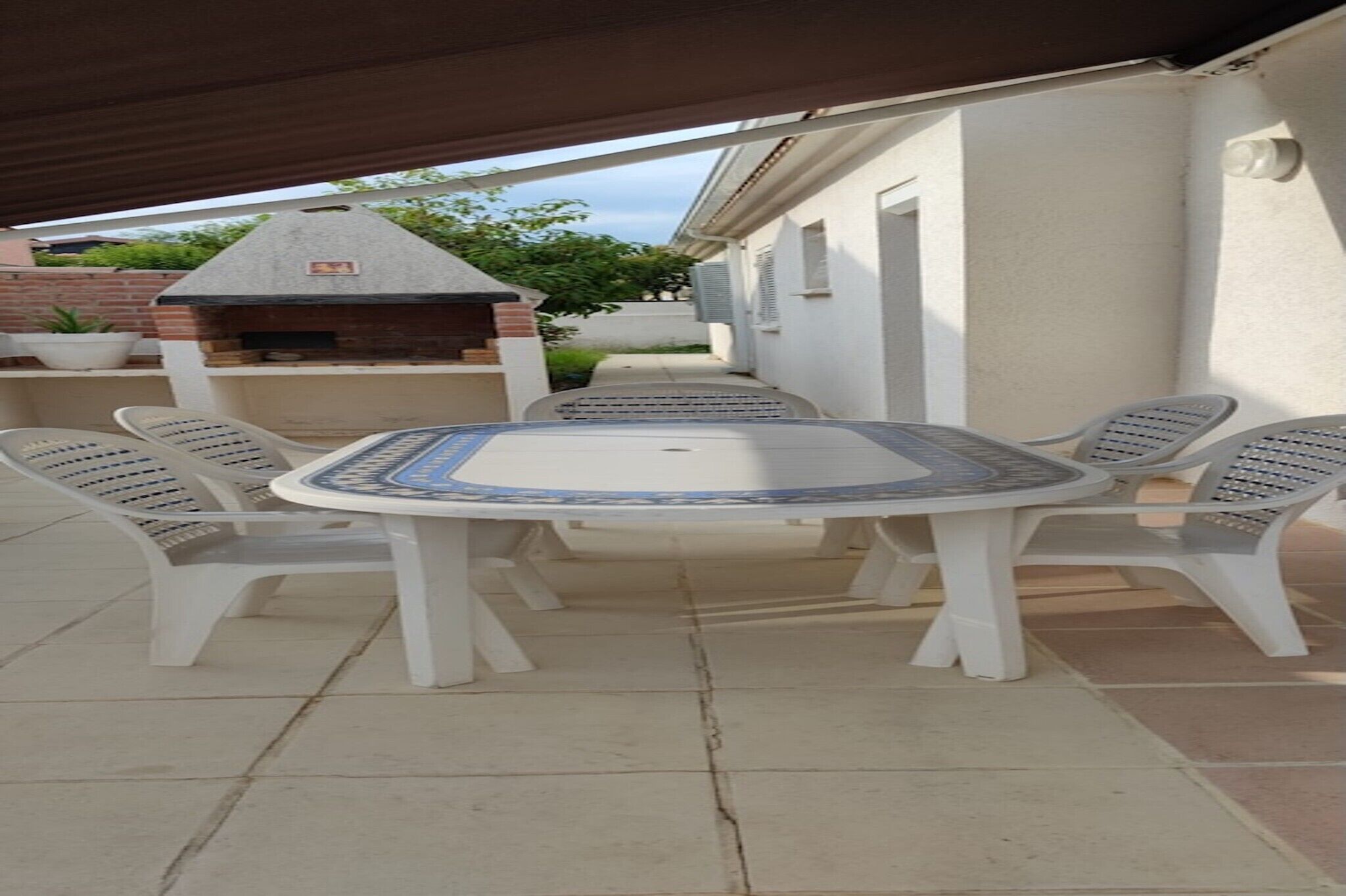 A covered patio area with a round table and four chairs, a brick barbecue grill, and a potted plant.