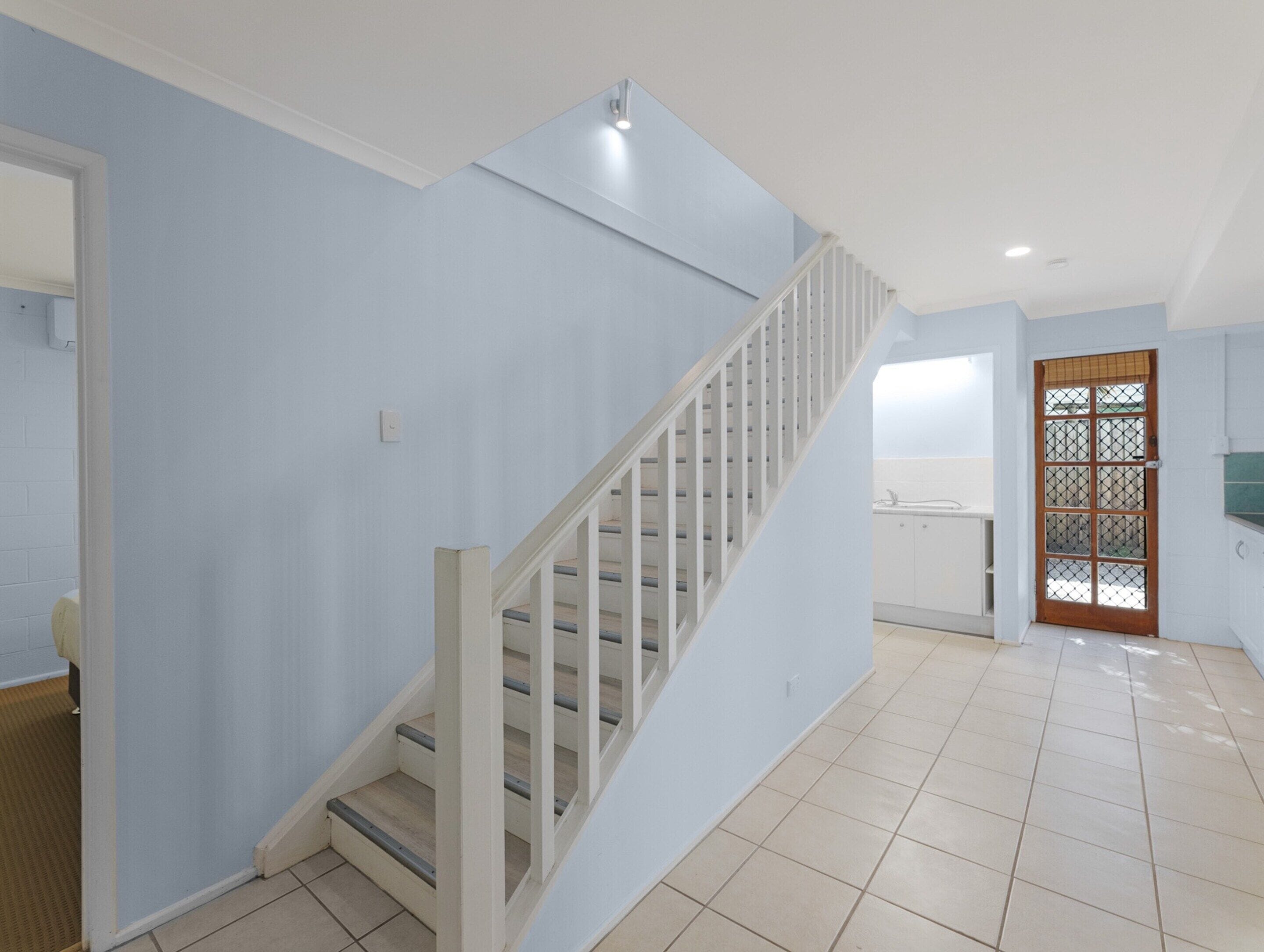 A spacious room with a staircase leading to an upper floor, a kitchen area, and a door with a security screen.