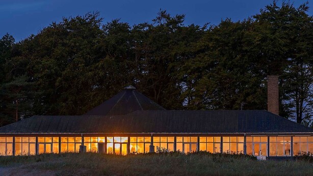 Un grand bâtiment d’un seul étage, doté d’un toit à pignon et d’une cheminée, entouré d’arbres denses.