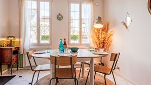 Ein Essbereich mit einem runden Tisch, Stühlen und einer Lampe. Es gibt Fenster mit Vorhängen, eine Uhr an der Wand und eine Heizung unter dem Tisch.