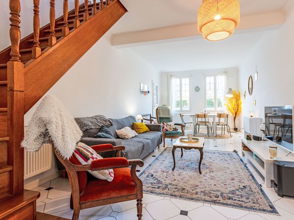 A spacious living room with a wooden staircase, a sofa, a red armchair, a coffee table, and a dining area with a table and chairs.