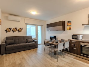 A modern living space with a sofa, dining table, chairs, a TV, kitchen area with cabinets, and a window. - Apartment Ilaria by Interhome (Riva del Garda)