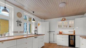 Une cuisine moderne avec des meubles blancs, des plans de travail en bois et un grand îlot central. De nombreuses fenêtres laissent entrer la lumière naturelle, et une horloge est visible au mur.