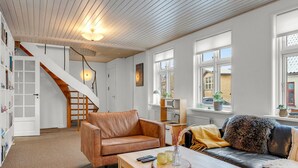 Un salon moderne avec un plafond en bois, un escalier, un canapé en cuir, une table basse en bois et une vue sur l’extérieur grâce à de grandes fenêtres.