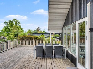 En træterrasse med udendørs møbler, en glasdør der fører til et rum, og udsigt til grønne områder og en bygning i baggrunden.