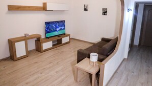 En moderne stue med trægulv, et fladskærms TV på et træstel, en brun sofa, et lille træbord med en lampe og en hvid væg med en loftlampe.