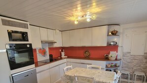 A compact kitchen with white cabinets, a built-in oven, a microwave, and a dishwasher. There is a dining table with chairs and a chandelier above.