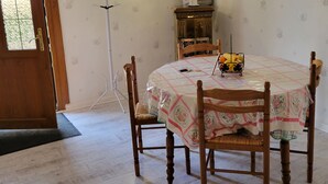 A dining area with a round table covered by a patterned tablecloth, surrounded by wooden chairs. A vintage-style lamp and a small decorative item are on the table. The room has patterned wallpaper and a wooden cabinet.