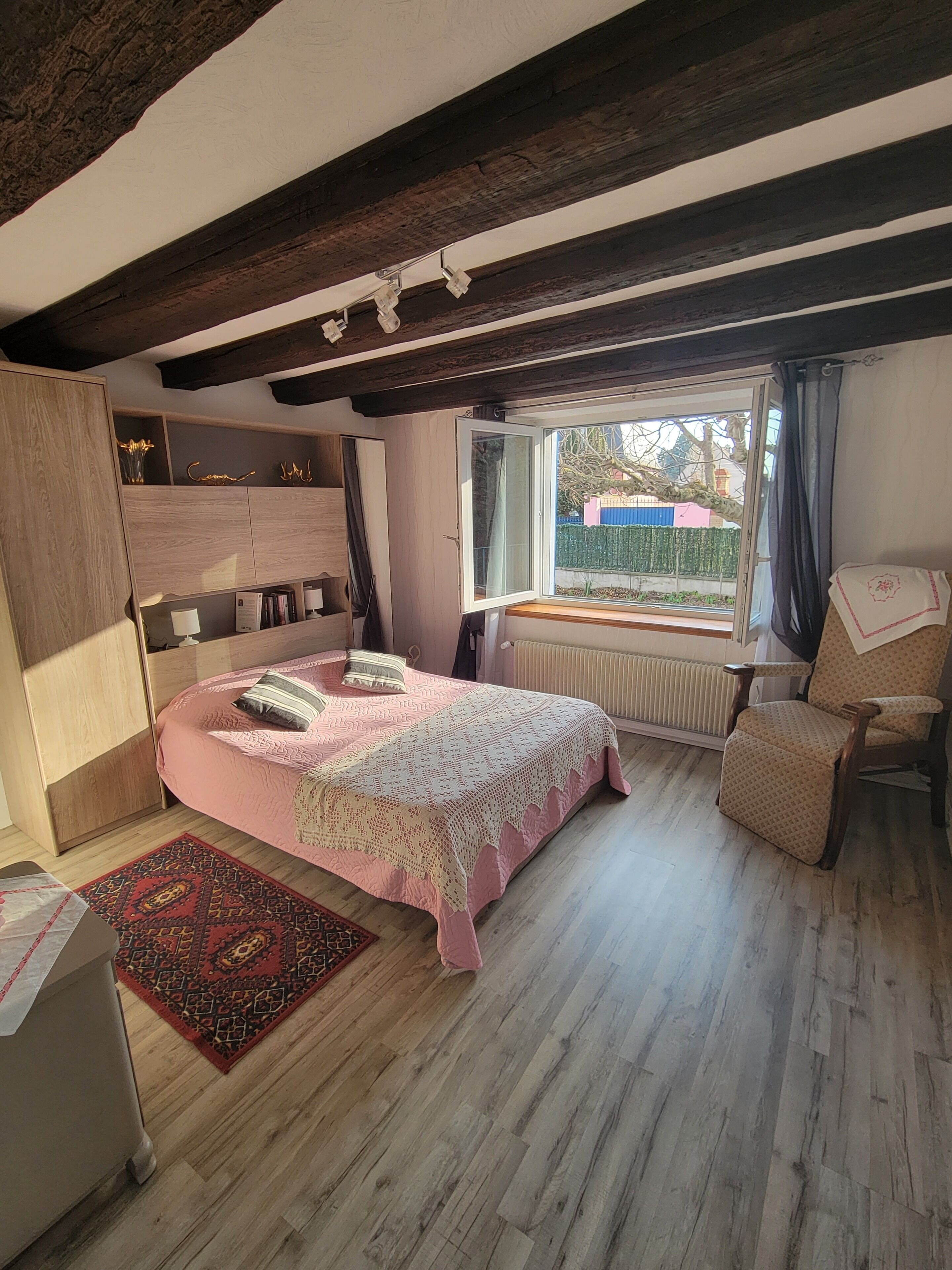 Un dormitorio con una cama de madera, un ventanal con vistas, un radiador y una silla.