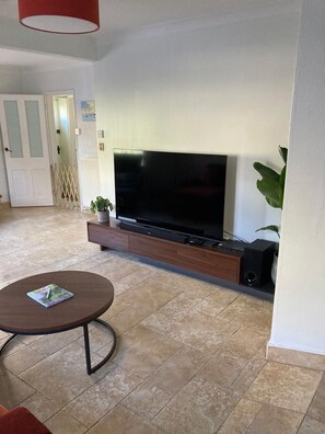 Ein Wohnzimmer mit einem großen Flachbildfernseher auf einem Holzständer, einem kleinen runden Tisch mit einer Zeitschrift und einer Topfpflanze.