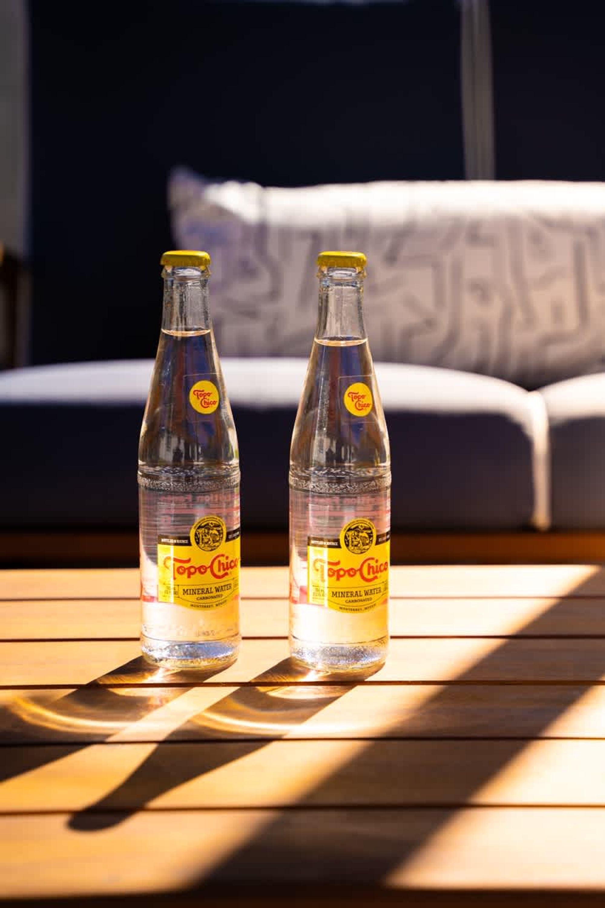 Two bottles of Topo Chico mineral water on a wooden table with sunlight casting shadows.