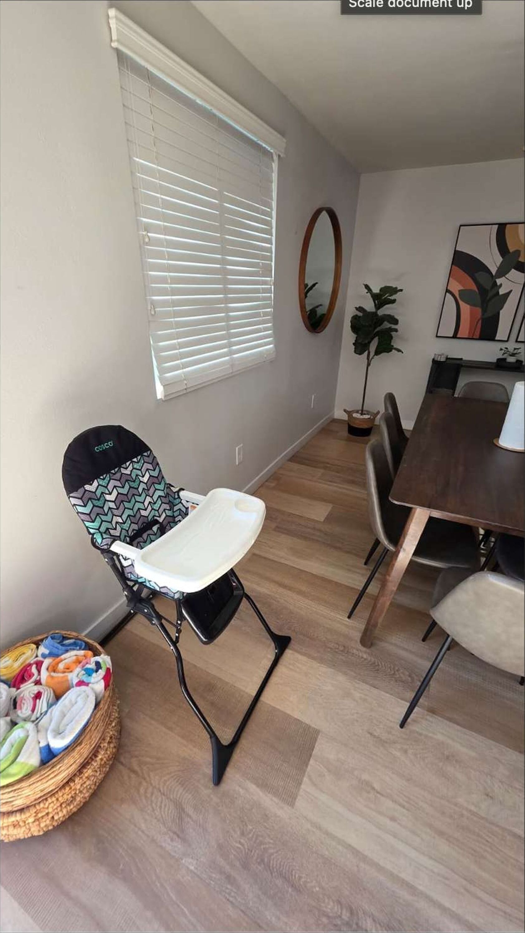 A room with a high chair, dining table, chairs, a potted plant, and a painting on the wall.
