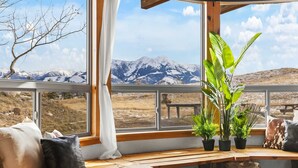 Eine gemütliche Sitzecke mit einer hölzernen Bank, Kissen und Blumentöpfen an einem Fenster mit Blick auf eine Berglandschaft.