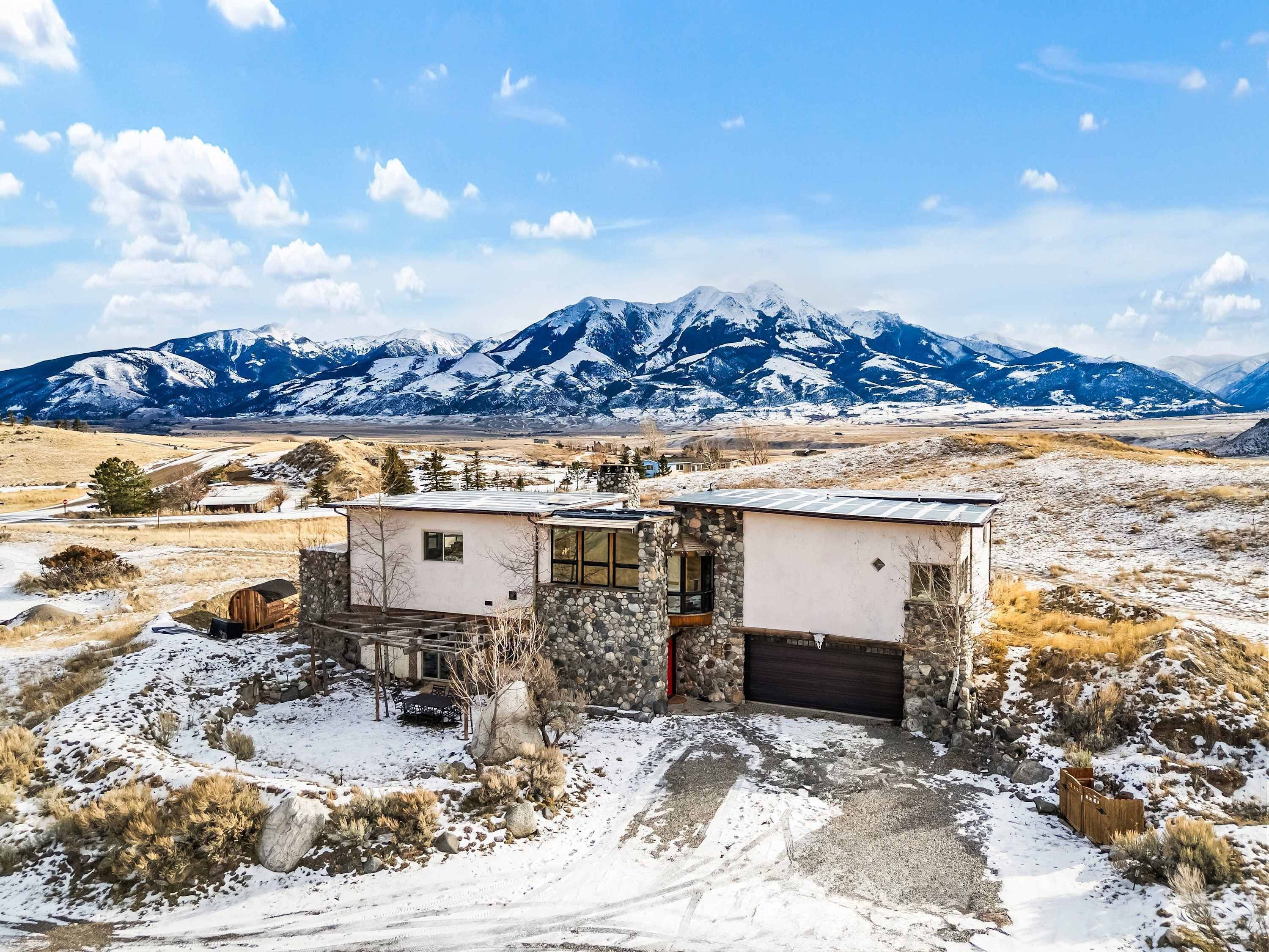 Una casa moderna con fachada de piedra, ventanas amplias y un garaje, con montañas nevadas y cielo despejado de fondo.