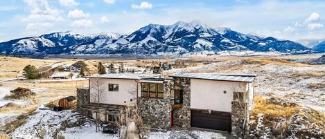 Una casa moderna con fachada de piedra, ventanas amplias y un garaje, con montañas nevadas y cielo despejado de fondo.