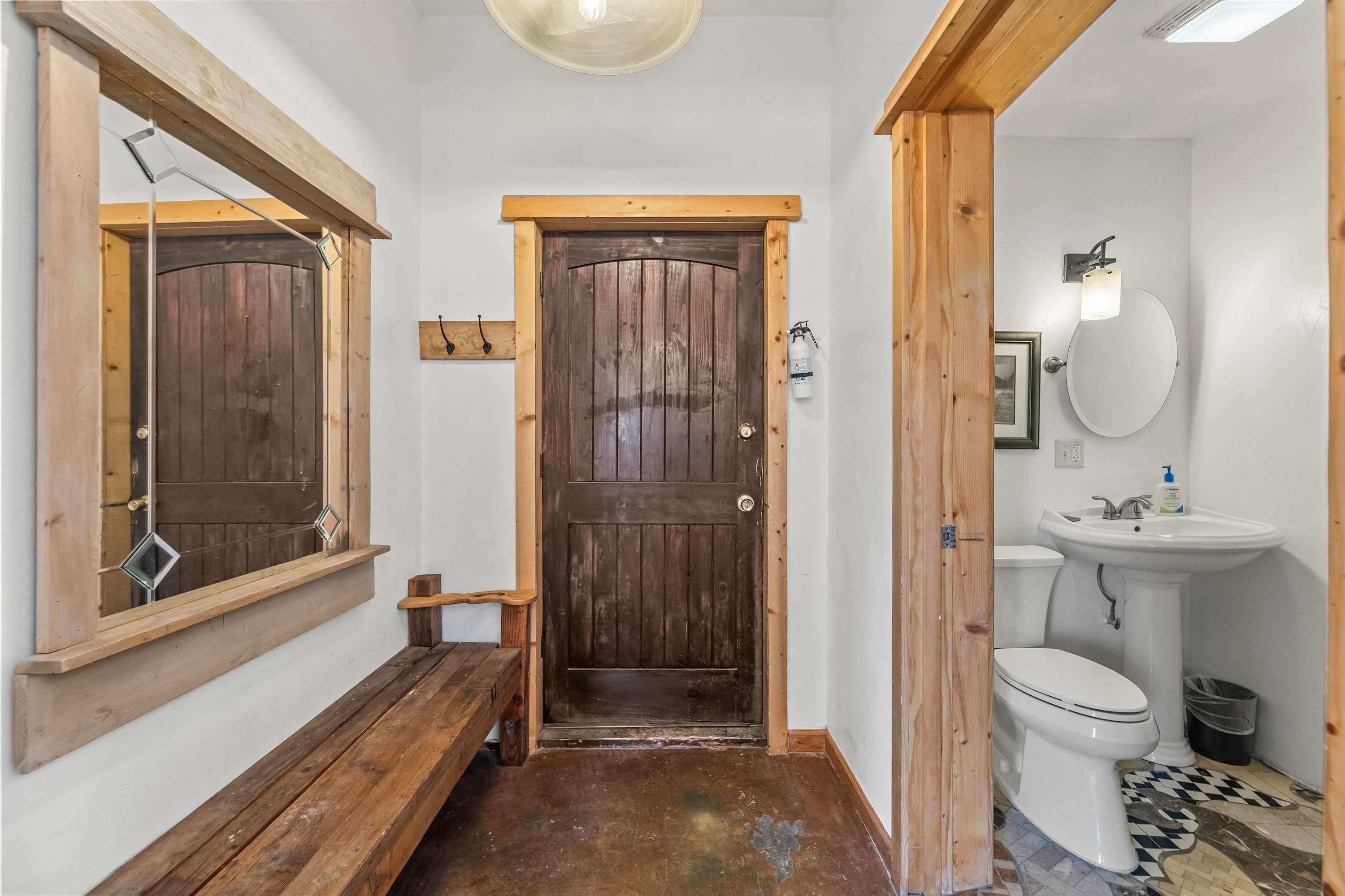 A wooden door with a mirror above it, a bench, a white sink, and a toilet in a small bathroom.