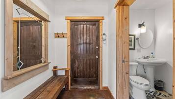 A wooden door with a mirror above it, a bench, a white sink, and a toilet in a small bathroom.