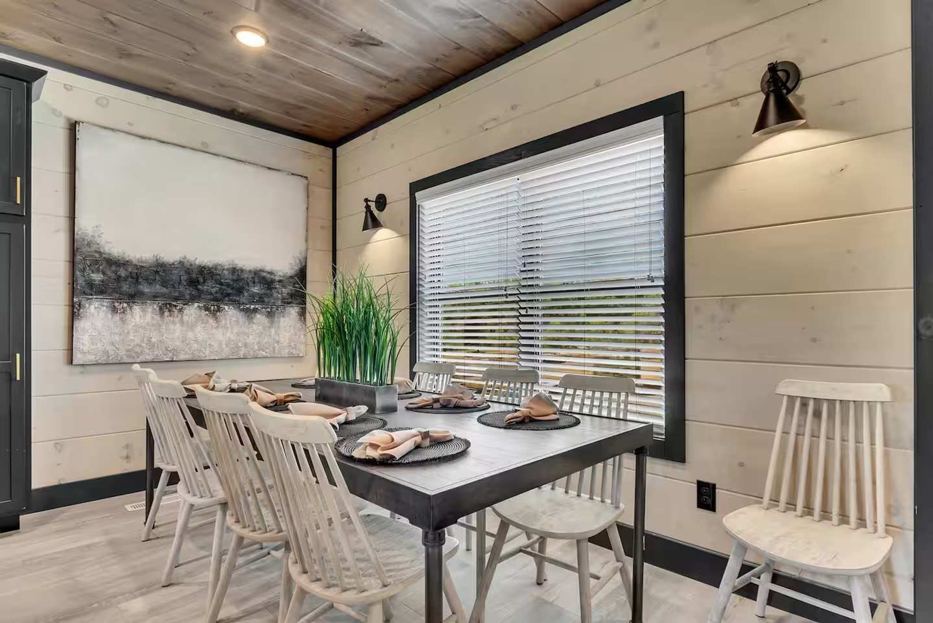 A dining area with a long table, white chairs, and a large window with blinds.