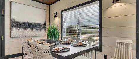 A dining area with a long table, white chairs, and a large window with blinds.