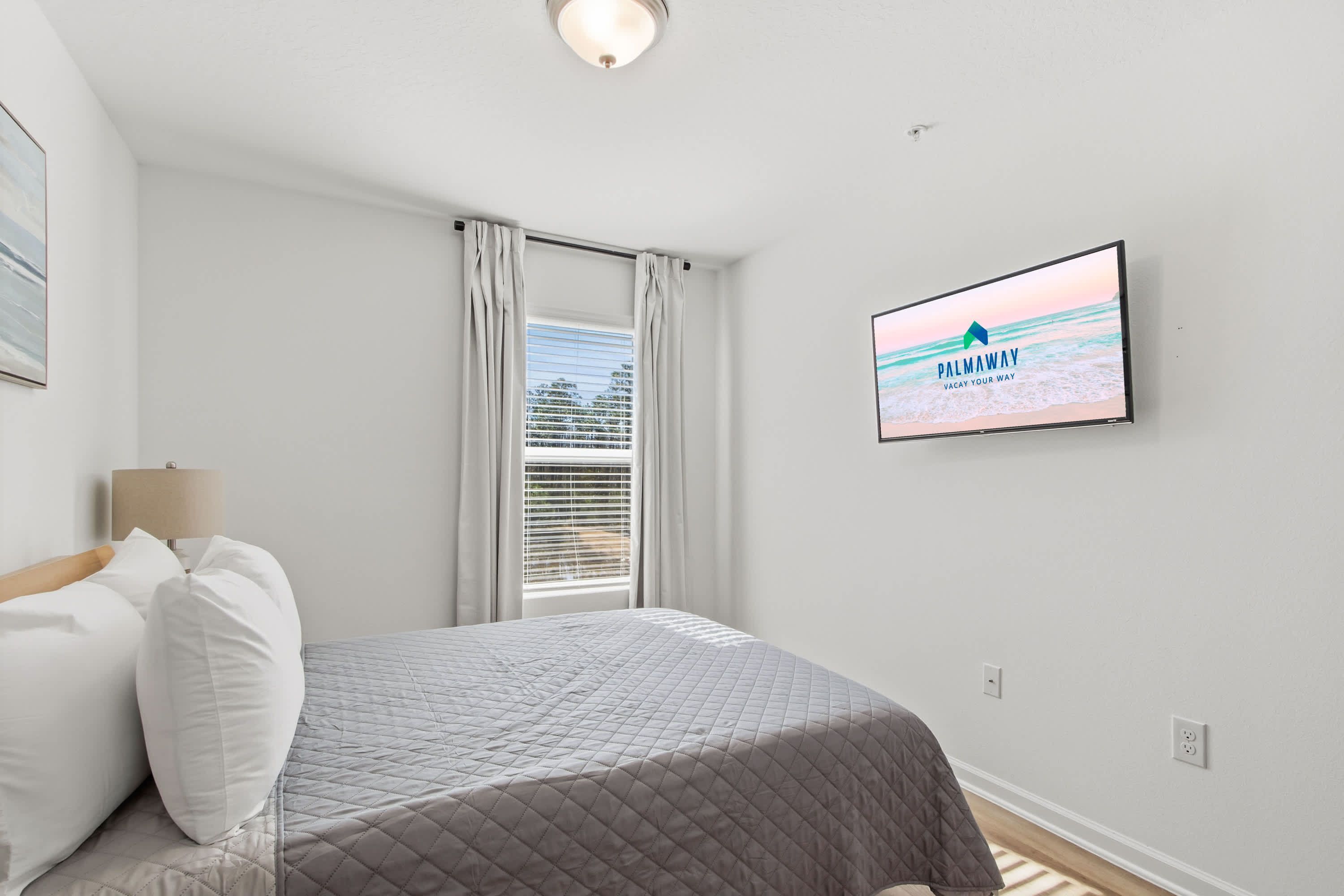 A bedroom with a bed, a TV mounted on the wall displaying 'PALMAWAY VACAY YOUR WAY', a window with curtains, and a ceiling light.