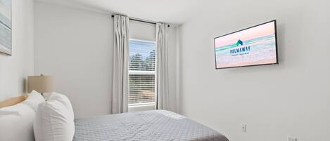 A bedroom with a bed, a TV mounted on the wall displaying 'PALMAWAY VACAY YOUR WAY', a window with curtains, and a ceiling light.
