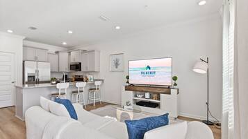 A modern living room with a white sofa, a flat-screen TV, and a kitchen in the background.