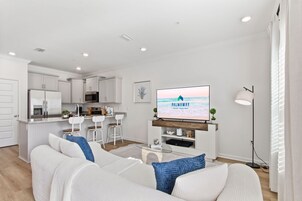 A modern living room with a white sofa, a flat-screen TV, and a kitchen in the background.