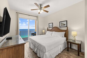 Een hotelkamer met een bed, een televisie, een plafondventilator, een balkon met uitzicht en ingekaderde foto's aan de muur.