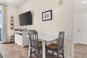 Een eetruimte met een witte tafel en twee stoelen, een flatscreen televisie aan de muur en een deur aan de rechterkant.