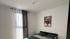 A bedroom with a bed, a nightstand, a radiator, a window with curtains, and a wall-mounted artwork. - New house or town center + parking space (Dijon)