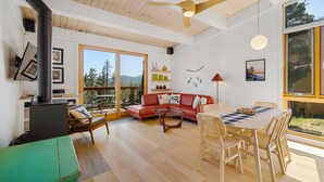 Un salón espacioso con techo de madera, un sofá rojo, una mesa de comedor con sillas y un ventanal con vistas a árboles y montañas.