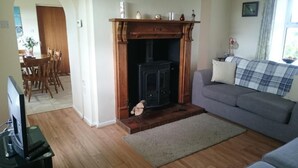 A living room with a fireplace, a grey sofa, a wooden dining table with chairs, and a television on a stand. - Portrush Country Cottage with sea views. Perfectly situated for sightseeing. (Coleraine)
