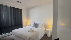 A modern bedroom with a large bed, two bedside tables with lamps, a window with blinds, and a door. - Innerbloom Stay Busselton - Guest House (West Busselton)