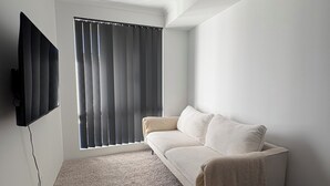 A minimalist living room with a large window, a flat-screen TV mounted on the wall, and a cozy sofa with a throw blanket. - Innerbloom Stay Busselton - Guest House (West Busselton)