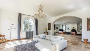 Ein großzügiger Wohnraum mit einem weißen Ecksofa, einer Chandelierleuchtreihe und Blick durch einen Rundbogen zur Küche.