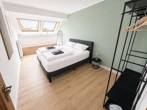 A bedroom with a bed, a small bedside table, a hanging rack, and a window with a view.