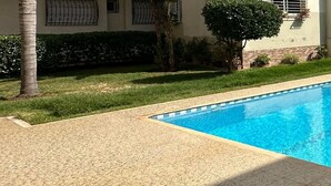 A rectangular swimming pool with clear blue water, surrounded by a tiled deck and a grassy area with small trees and shrubs. - Résidence Luxueuse, 2 Piscines Escapade Familiale à 2 pas Plages Harhoura Rabat (Harhoura)