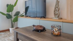 Ein modernes Wohnzimmer mit einem Flachbildfernseher auf einem Holzregal, einer Topfpflanze, einer dekorativen Schildkröte und einem Vogelkäfig mit Verzierungen.