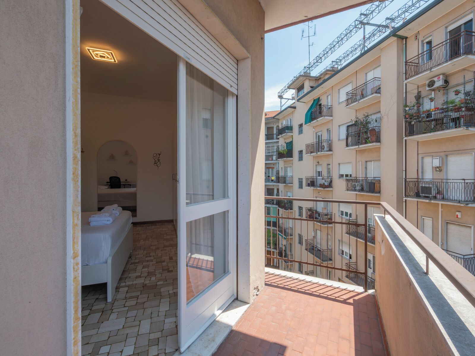 Un balcon avec un sol carrelé, une porte blanche donnant sur une pièce avec un lit et une petite table, et une vue sur un immeuble à plusieurs étages doté de balcons.