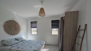 A bedroom with a bed, two windows with blinds, a woven wall hanging, a wooden ladder, and a ceiling light. - Quiet new villa - Tropical relaxation guaranteed (Saint-François)