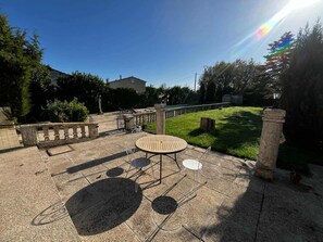 Een terras buiten met een ronde tafel en stoelen, een stenen balustrade en een gazon.