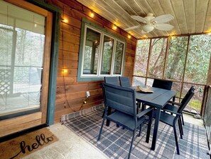 Eine Holzhütte mit Essbereich, inklusive Tisch und Stühlen, Deckenventilator und Blick auf Bäume durch große Fenster.