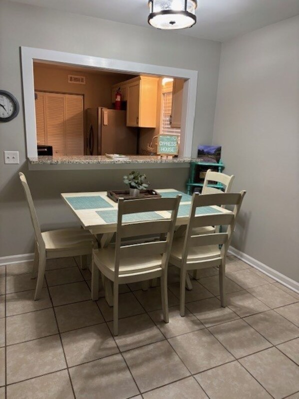 A dining area with a table and four chairs, a clock on the wall, and a kitchen visible through an open door.