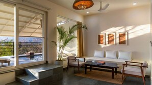 A modern living room with a sofa, coffee table, and chairs, large windows with a view of a pool and greenery, and a ceiling fan. - Vihara Udaipur (Udaipur)