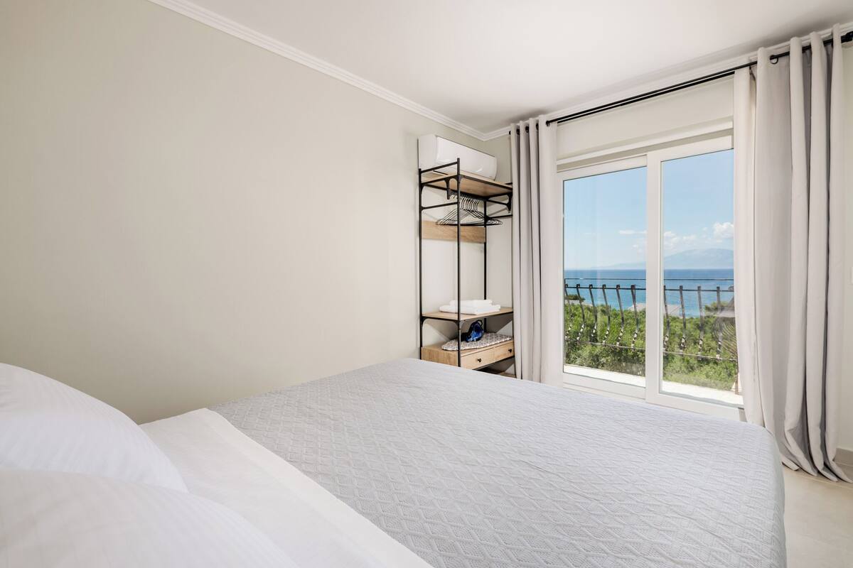 A hotel room with a bed, a built-in shelf with a mirror, and a large window offering a view of the sea.