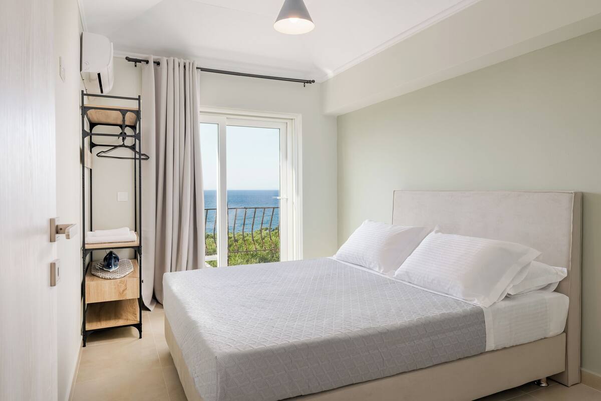 A bedroom with a large bed, a wardrobe, a small table, and a window with a view of the sea.