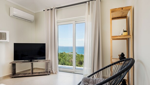 A modern living room with a large flat-screen TV, a wicker chair, and a view of the ocean through floor-to-ceiling windows. - Restia tower apartments (Zakynthos)