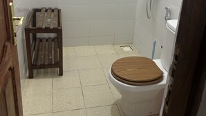 A small, tiled bathroom with a toilet, a wooden stool, and a shower. - Spacious Deluxe Double Room With AC (Paje)