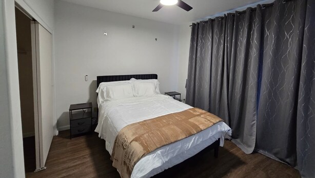 Une chambre à coucher avec un lit, une table de chevet, un ventilateur de plafond et une fenêtre avec des rideaux.